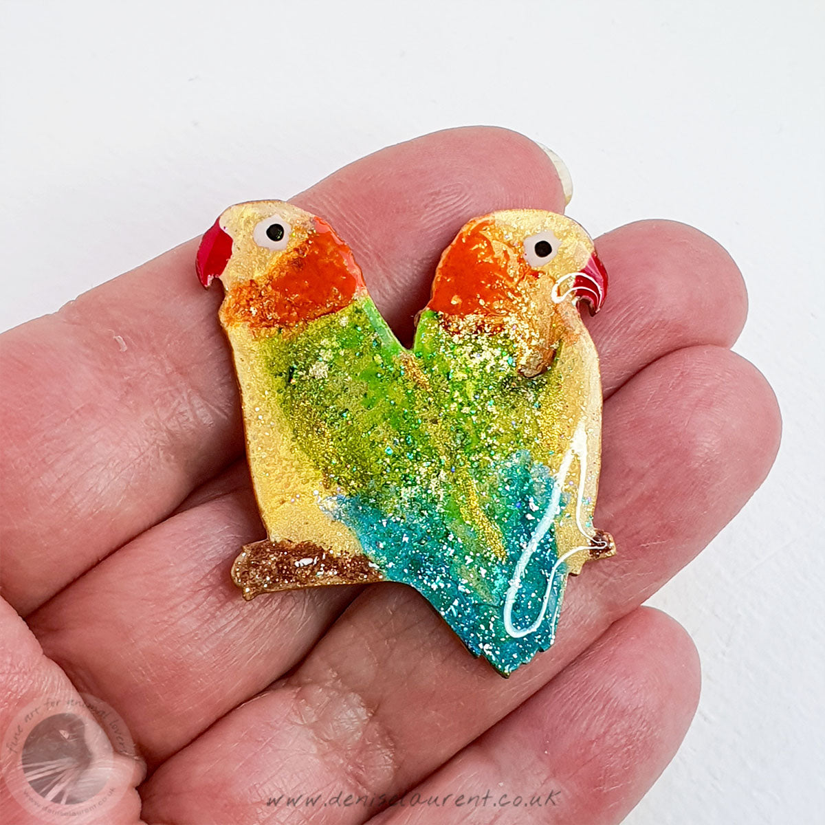 A hand holding a bird pin. A pair of Lovebirds. They have yellow and orange heads with green and blue wings. They have touches of brilliant gold leaf through them.