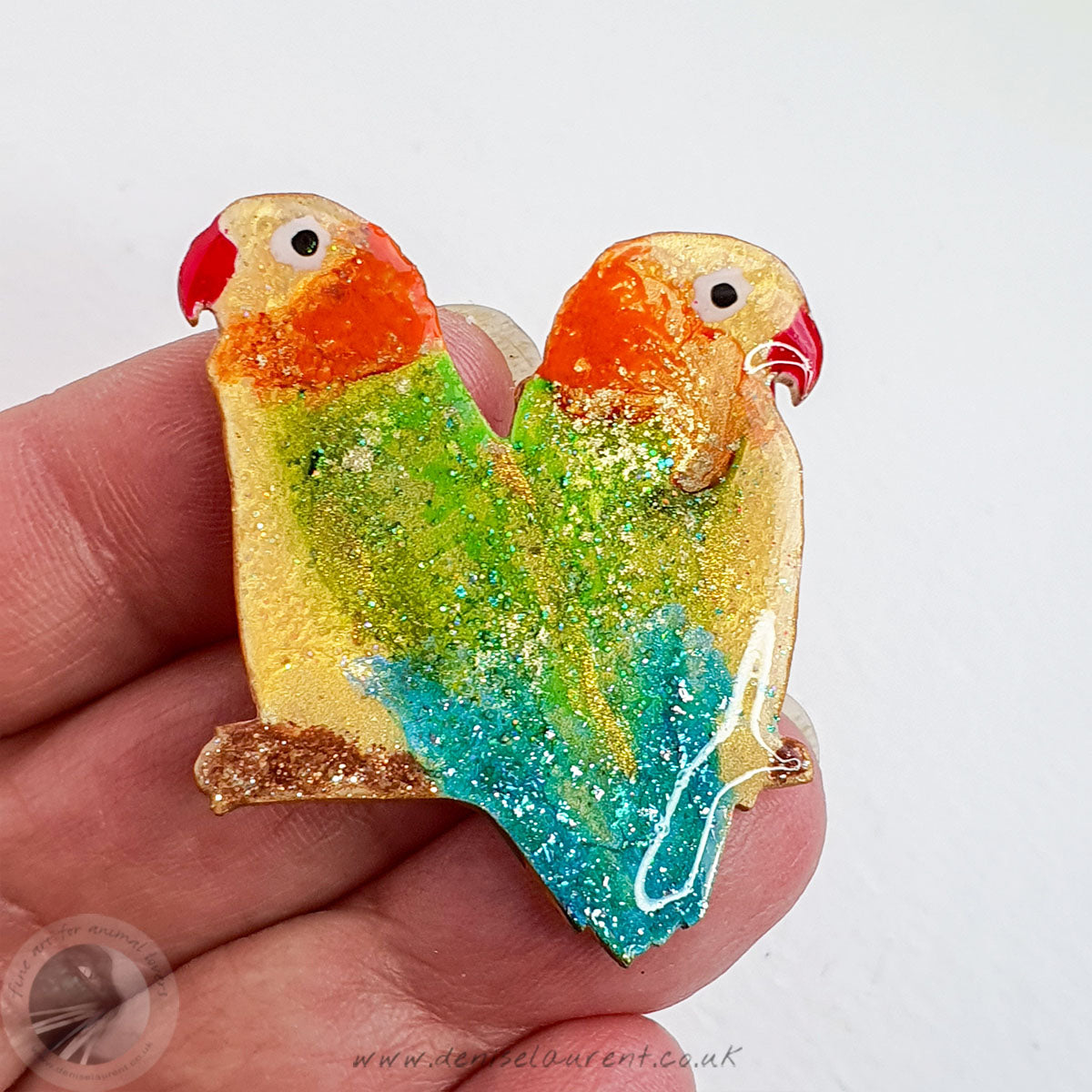 A hand holding a lovebird brooch. They have yellow and orange heads with green and blue wings. They have touches of brilliant gold leaf through them.