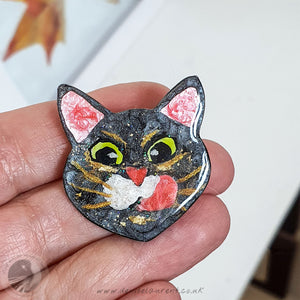 A handmade, one-of-a-kind brooch of black cat face, with big green eyes, white whisker pads and gold whiskers, licking his lips, balanced on a hand.