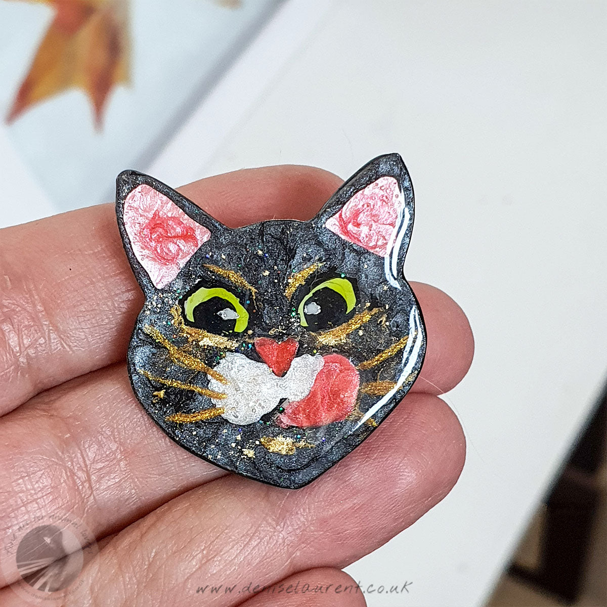 A handmade, one-of-a-kind brooch of black cat face, with big green eyes, white whisker pads and gold whiskers, licking his lips, balanced on a hand.