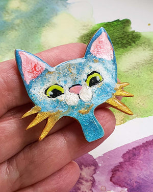 a hand holding  blue cat face brooch with a big smile and gold whiskers.