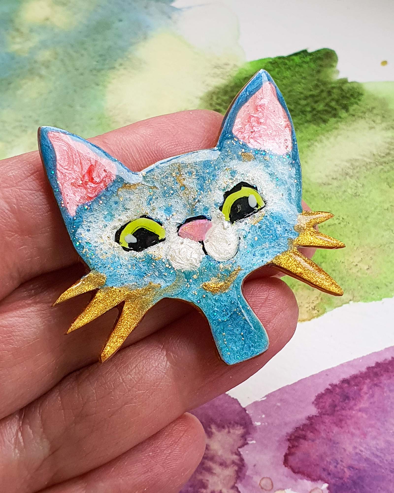 a hand holding  blue cat face brooch with a big smile and gold whiskers.