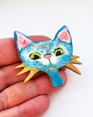 A hand holding a blue, white and gold kitty pin. This cute broach has gold whiskers, big green eyes and a pink nose.