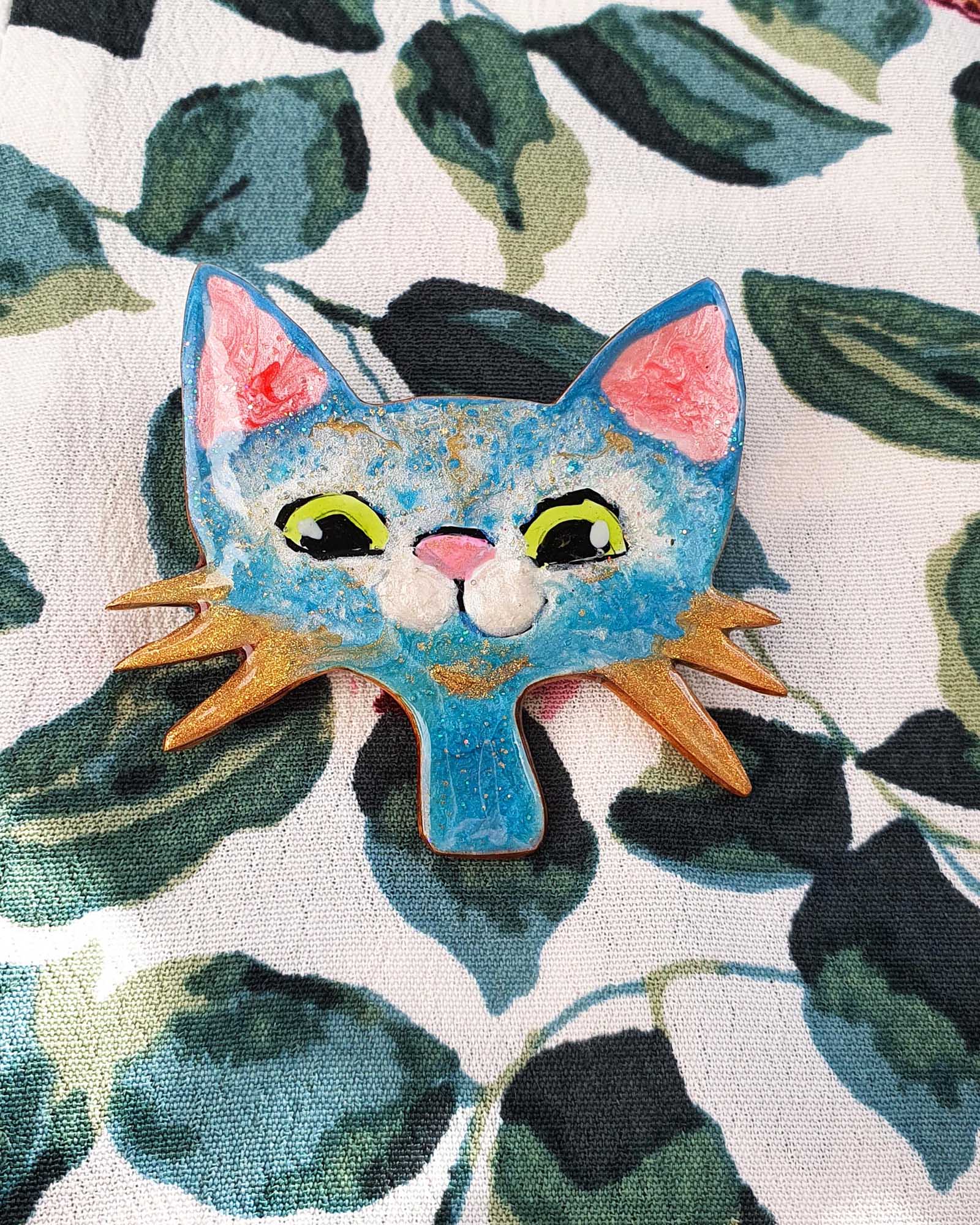 A blue, white and gold cat broach on a fabric top.  This whimsical cat brooch has gold whiskers, big green eyes and a pink nose.