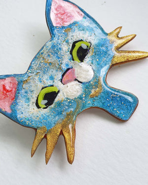 A close up detail of a blue, white and gold cat brooch on a white background. This cute brooch has gold whiskers, big green eyes and a pink nose. You can see the glitter of the gold leaf.