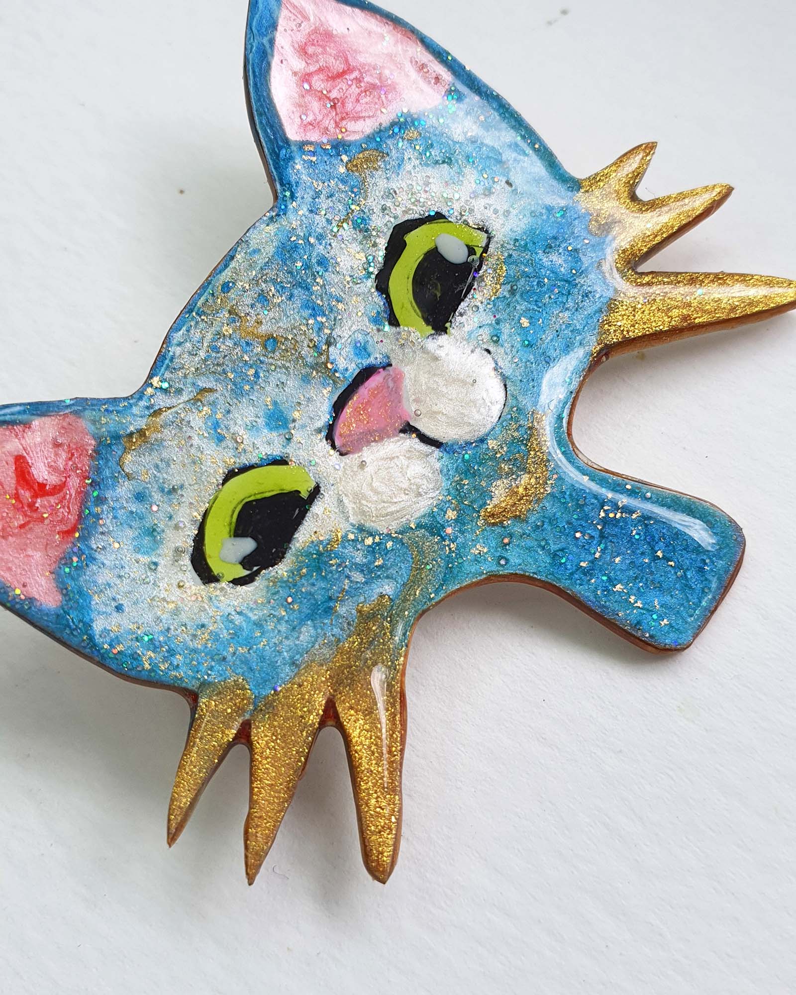 A close up detail of a blue, white and gold cat brooch on a white background. This cute brooch has gold whiskers, big green eyes and a pink nose. You can see the glitter of the gold leaf.