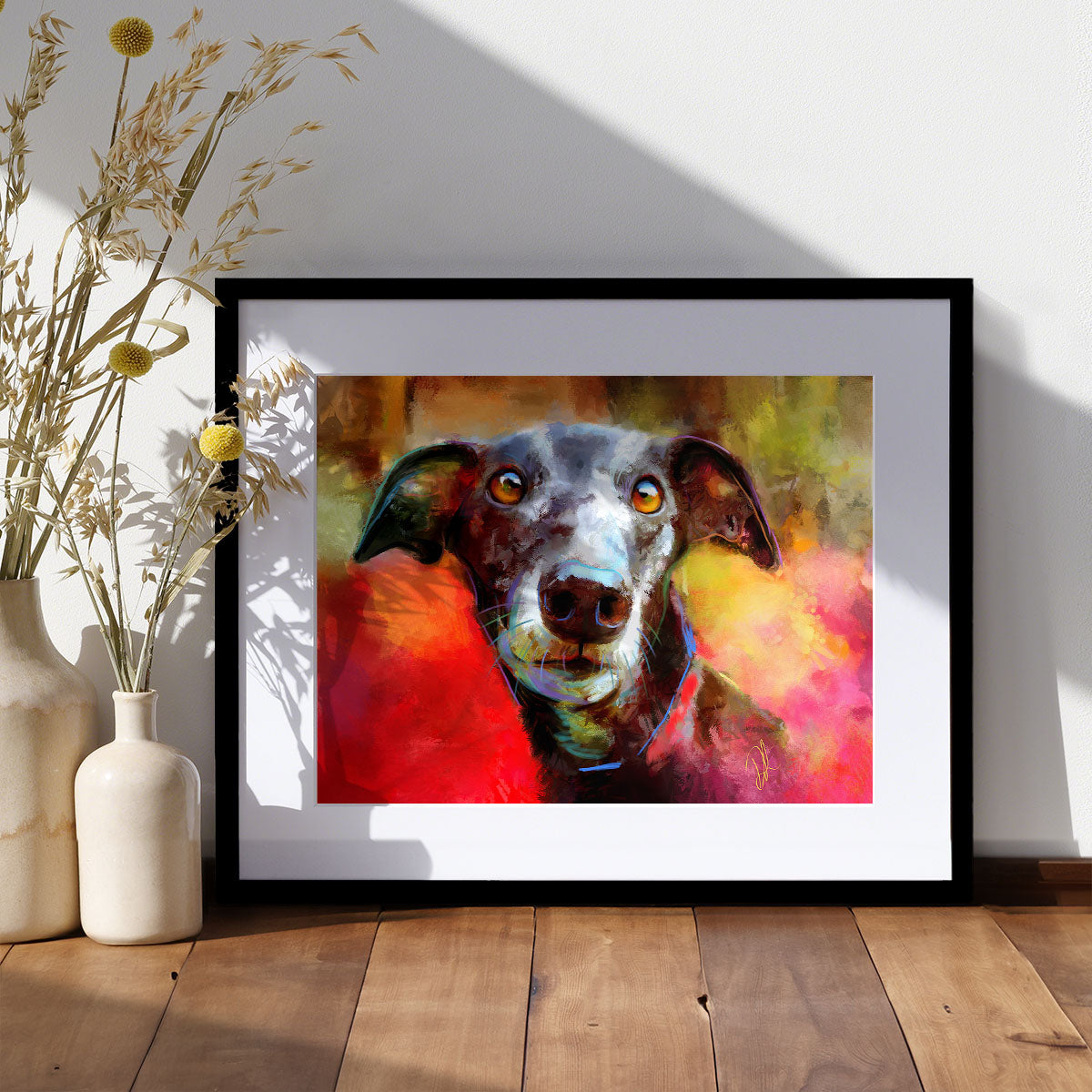 A framed and mounted print on paper of a black dog on a colourful background of red and golds. The framed print sits on a wooden floor.