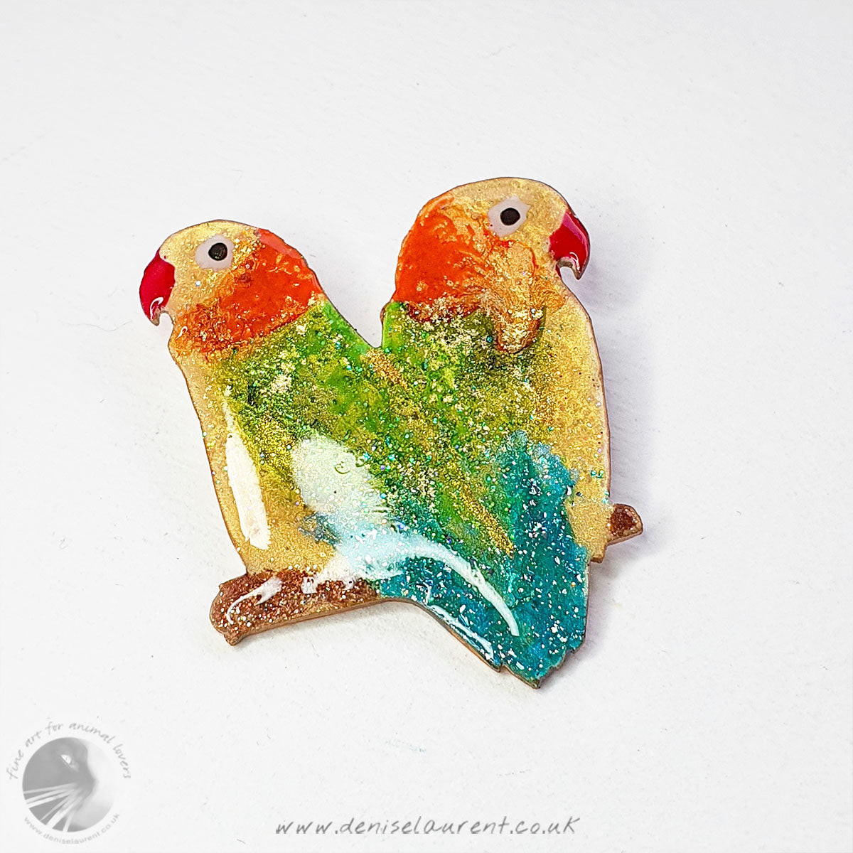 A hand holding a handmade lovebird brooch, this broach has a pair of lovebirds sitting on a branch back to back. They are yellow, green, orange and blue. 