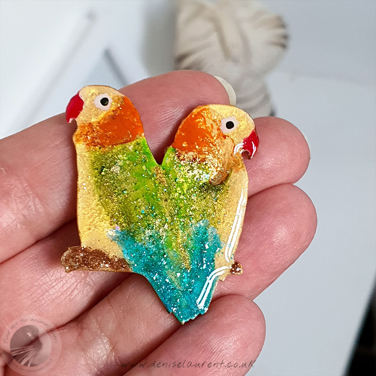 A hand holding a handmade lovebird brooch, this broach has a pair of lovebirds sitting on a branch back to back. They are yellow, green, orange and blue. 