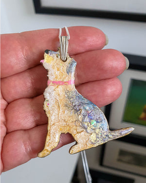 Border terrier dog-shaped necklace. One side of a double sided pendant. This side has the colours of a border terrier with a pink colour and a pink tongue. held in a hand with a blurred background