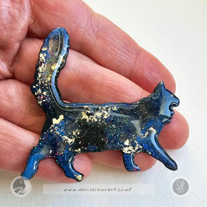 Blue and gold cat brooch held in a hand on a white background