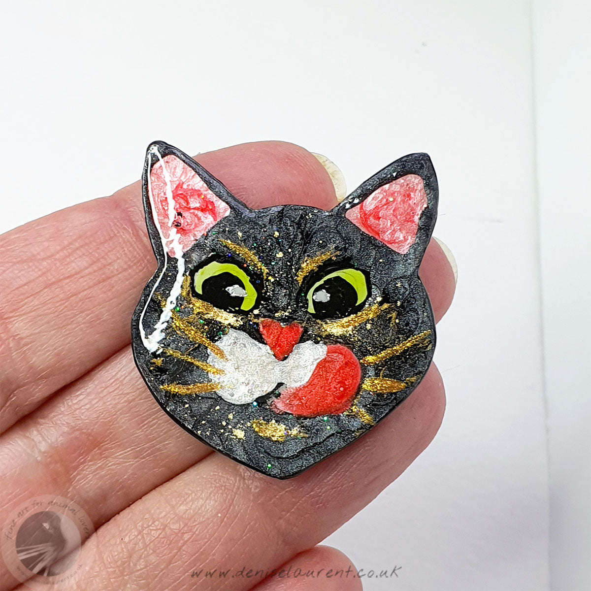A cute cat face brooch. A black cat face with big green eyes and gold whiskers, he is licking his lips.
