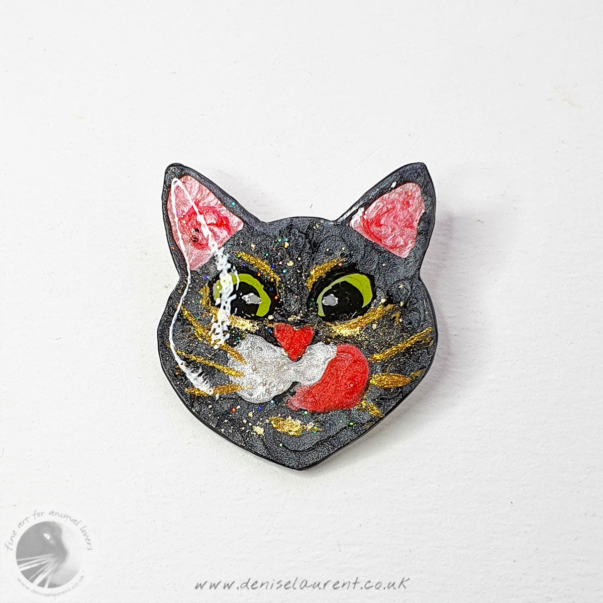 A cute cat face brooch. A black cat face with big green eyes and gold whiskers, he is licking his lips.