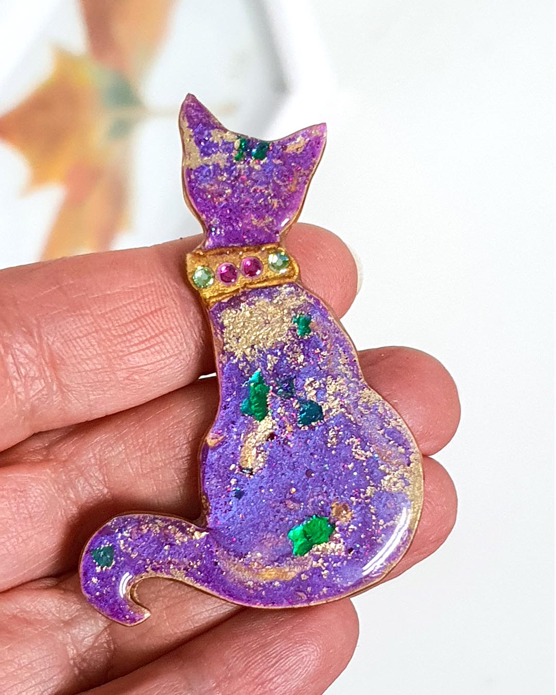 A purple and gold handmade cat brooch in the hand.