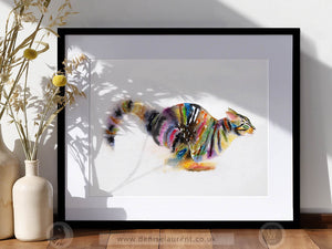 A framed watercolor art print of a colorful tabby cat, displayed in a black frame on a wooden surface next to decorative ceramic vases with dried yellow flowers. The print features a vibrant multicolor cat on a white background with soft leaf shadows cast across it.