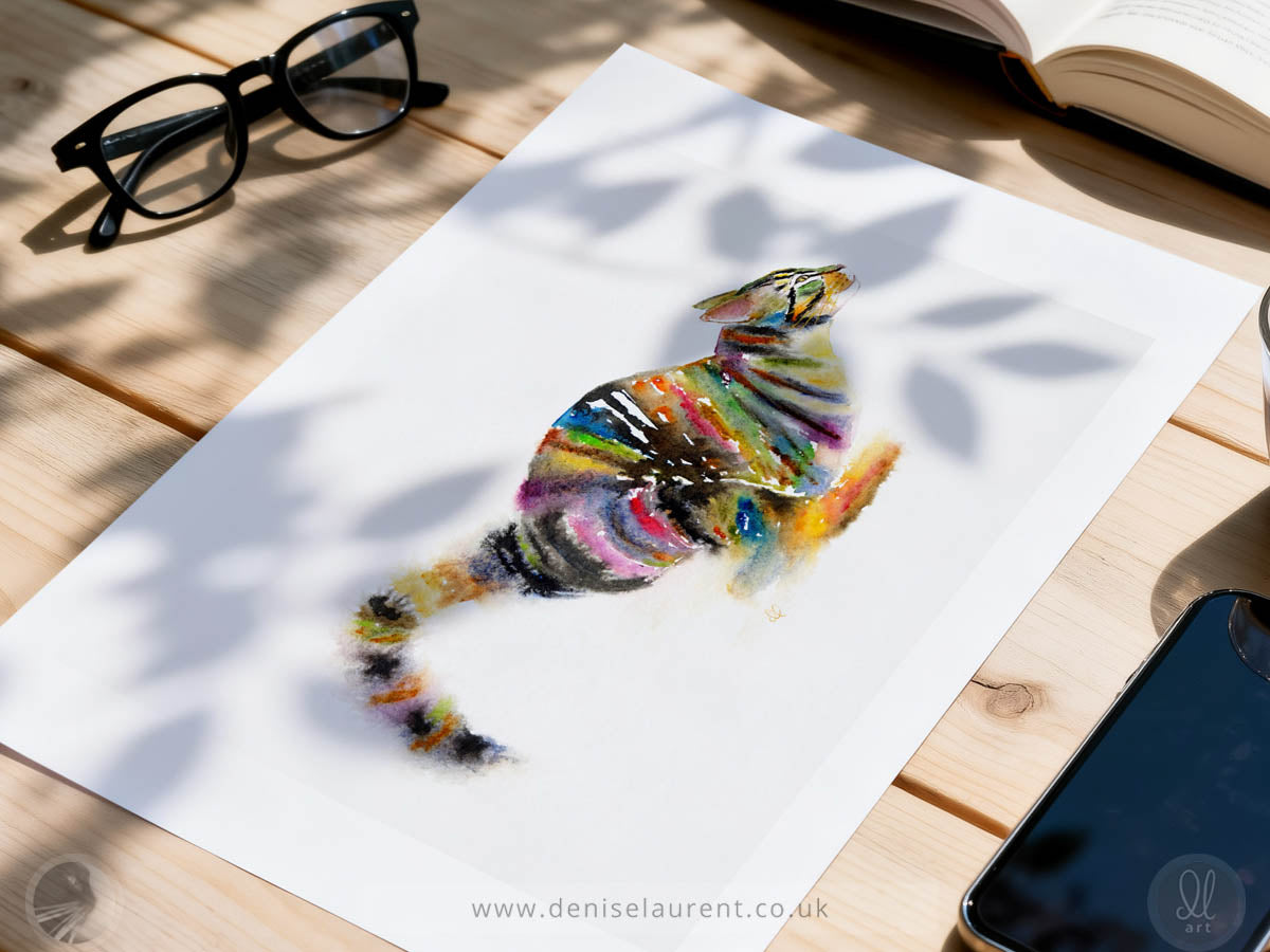 A white art print featuring a colorful watercolor-style cat illustration, displayed on a light wooden table with glasses, an open book, and a smartphone nearby.