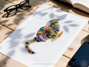 A white art print featuring a colorful watercolor-style cat illustration, displayed on a light wooden table with glasses, an open book, and a smartphone nearby.