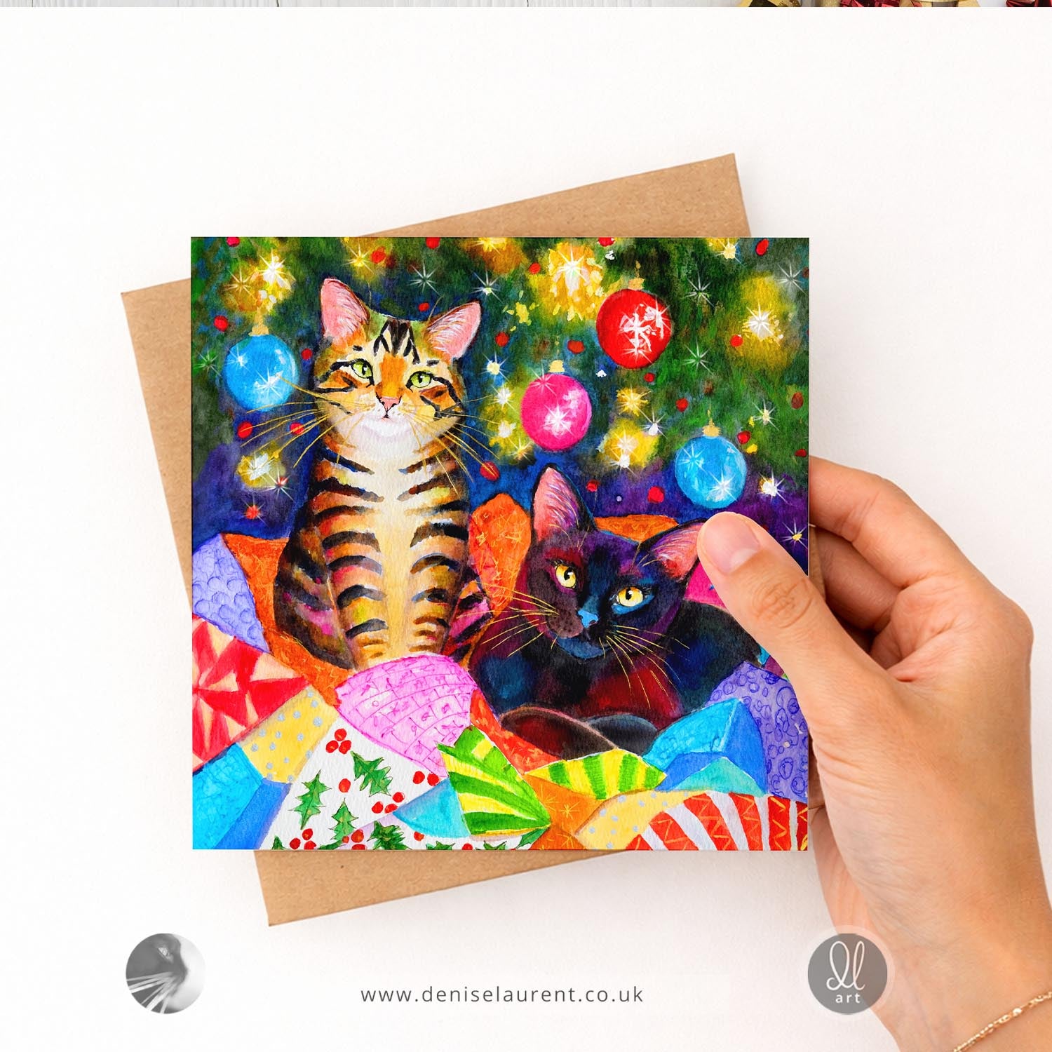 A square greeting card featuring a colorful painting of two cats in a festive Christmas scene, displayed on a windowsill with holiday decorations and blurred lights in the background.